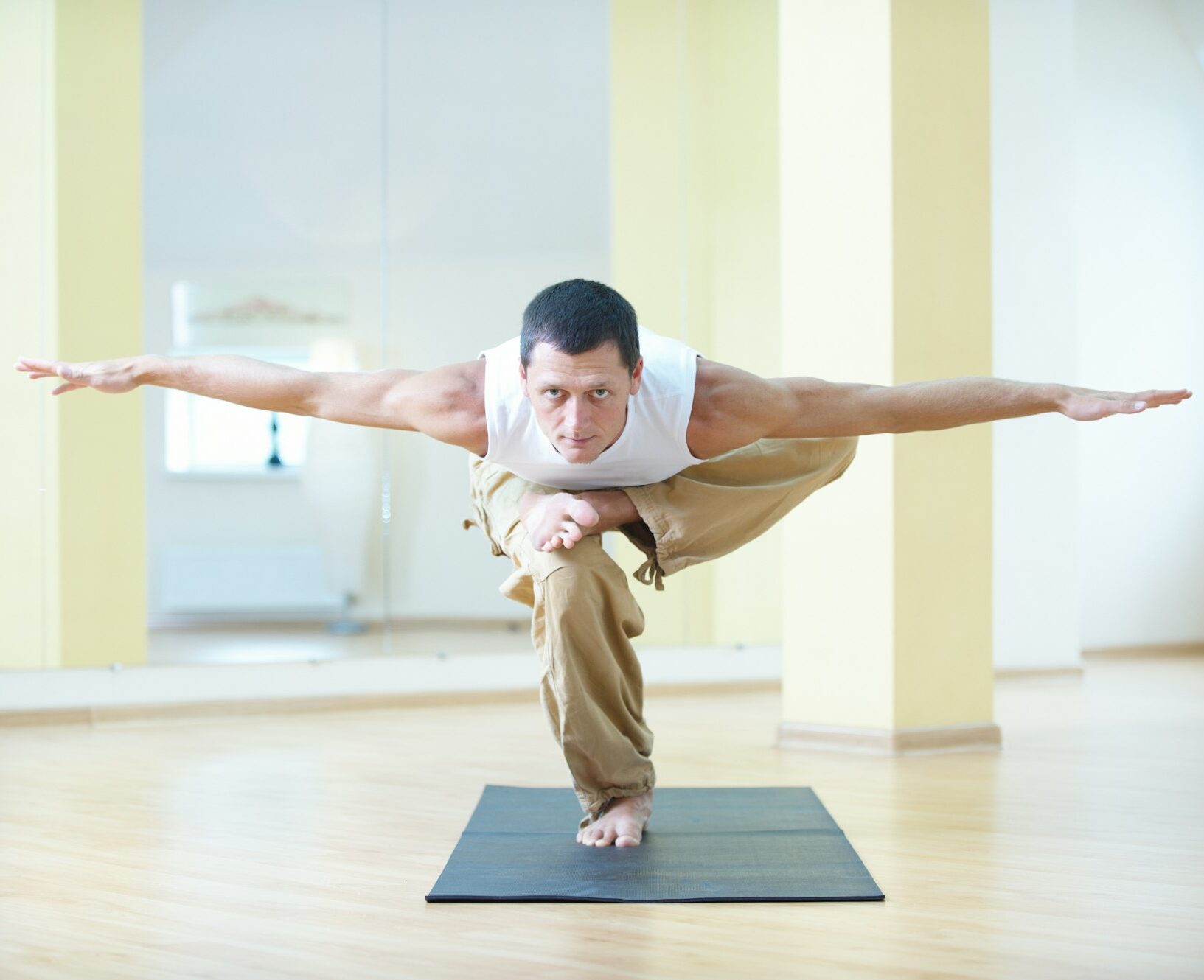 A young strong man stands in the asana on one leg with the arms apart in the yoga studio